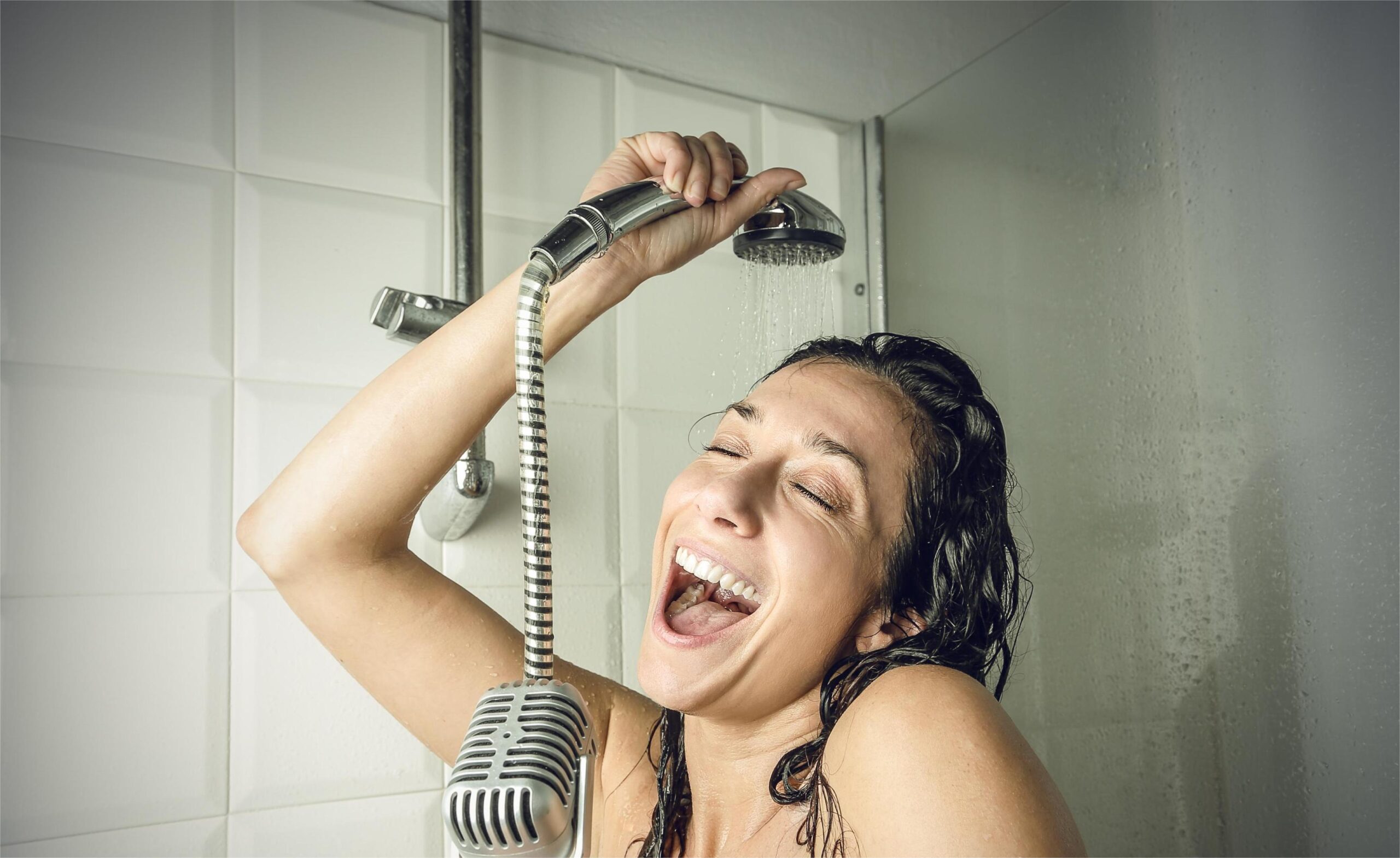 A woman singing in the shower with a shower head over her and a microphone.